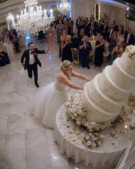 As I lifted the knife to cut the wedding cake, my sister hugged me tightly and whispered, “Push it over. Now.” I glanced at her, then at my smiling groom. Without thinking, I slammed the cake cart, sending the entire three-tier cake crashing to the floor as guests screamed. In the chaos, my sister grabbed my wrist and dragged me toward the side exit. “Run,” she hissed, her face pale. “You have no idea what he planned for you tonight.”